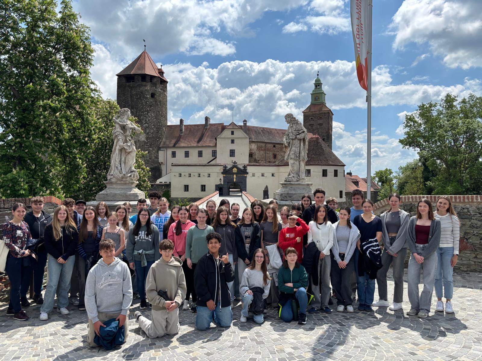 Das Bild zeigt eine Gruppe junger Menschen, die vor einer historischen Burganlage posieren. Im Hintergrund ist eine beeindruckende mittelalterliche Burg mit einem markanten Turm, weißen Mauern und roten Dächern zu sehen. Die Burg verfügt über einen Eingangsbereich mit Torbogen und ist von zwei Steinskulpturen flankiert.  Der Himmel ist strahlend blau mit weißen Wolken, was auf einen sonnigen Tag hindeutet. Rechts im Bild ist eine Fahne zu erkennen. Die Gruppe steht auf einem gepflasterten Vorplatz, einige Personen knien in der vorderen Reihe. Die jungen Leute tragen überwiegend Freizeitkleidung wie Jeans, T-Shirts und Sweatshirts in verschiedenen Farben.  Das Bild wirkt wie ein Gruppenausflug oder eine Schulexkursion zu dieser historischen Sehenswürdigkeit.