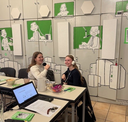 Bildbeschreibung  In einem hellen Raum sitzen zwei junge Personen an einem rechteckigen Tisch und lächeln in die Kamera. Beide halten jeweils ein kleines Mikrofon; eine Person trägt zusätzlich weiße Kopfhörer. Auf dem Tisch stehen ein Laptop mit geöffneter Präsentationsfolie, ein Wasserkrug, Gläser, Schälchen mit Snacks (u. a. Brotchips und geschnittenes Gemüse) sowie einzelne Notizzettel und Stifte.  Im Hintergrund ist eine Wand mit grauen Schranktüren zu sehen, die mit großflächigen schwarz-weißen Zeichnungen von Stadtgebäuden und stilisierten Figuren in grünen Fenstern gestaltet ist. Einige akustische Elemente (weiße, sechseckige oder rechteckige Paneele) sind an der Wand befestigt. Die Atmosphäre wirkt freundlich und kreativ, als handele es sich um einen Workshop oder eine Aufnahme-/Mediensession in einem Gemeinschafts- oder Unterrichtsraum.
