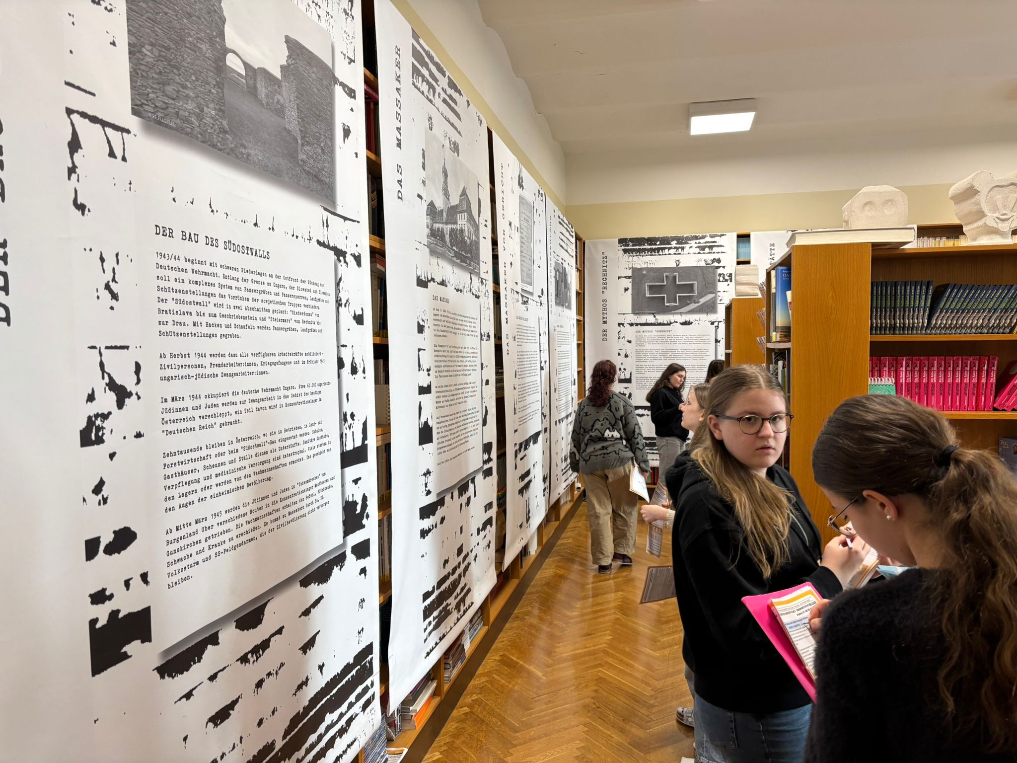 Das Bild zeigt einen Raum, der wie eine **Ausstellung oder ein Informationsbereich in einer Bibliothek oder Schule** wirkt. Entlang der linken Seite stehen große, weiße Ausstellungstafeln mit **schwarz-weißen Fotos und längeren Texten auf Deutsch**. Die Tafeln sind hoch und hängen dicht nebeneinander. Auf ihnen sind historische Bilder, Überschriften und erklärende Texte zu sehen, die offenbar ein **historisches Thema** behandeln.  Mehrere **Jugendliche bzw. Schülerinnen** stehen im Raum und betrachten die Ausstellung. Einige lesen die Tafeln aufmerksam, während zwei Personen im Vordergrund **Notizen auf einem Blatt oder in einer Mappe** machen. Sie wirken konzentriert und beschäftigen sich mit den Informationen der Ausstellung.  Der Raum hat einen **Holzboden im Fischgrätmuster** und auf der rechten Seite stehen **Bücherregale**, die mit Büchern gefüllt sind. Auf einem Regal liegen auch **Steinskulpturen oder Gipsmodelle von Köpfen**. Die Beleuchtung ist hell und kommt von einer Deckenlampe.  Insgesamt vermittelt das Bild den Eindruck eines **Lern- oder Bildungsprojekts**, bei dem Schülerinnen und Schüler eine **historische Ausstellung lesen und bearbeiten**, vermutlich im Rahmen von Unterricht oder einer Projektarbeit.