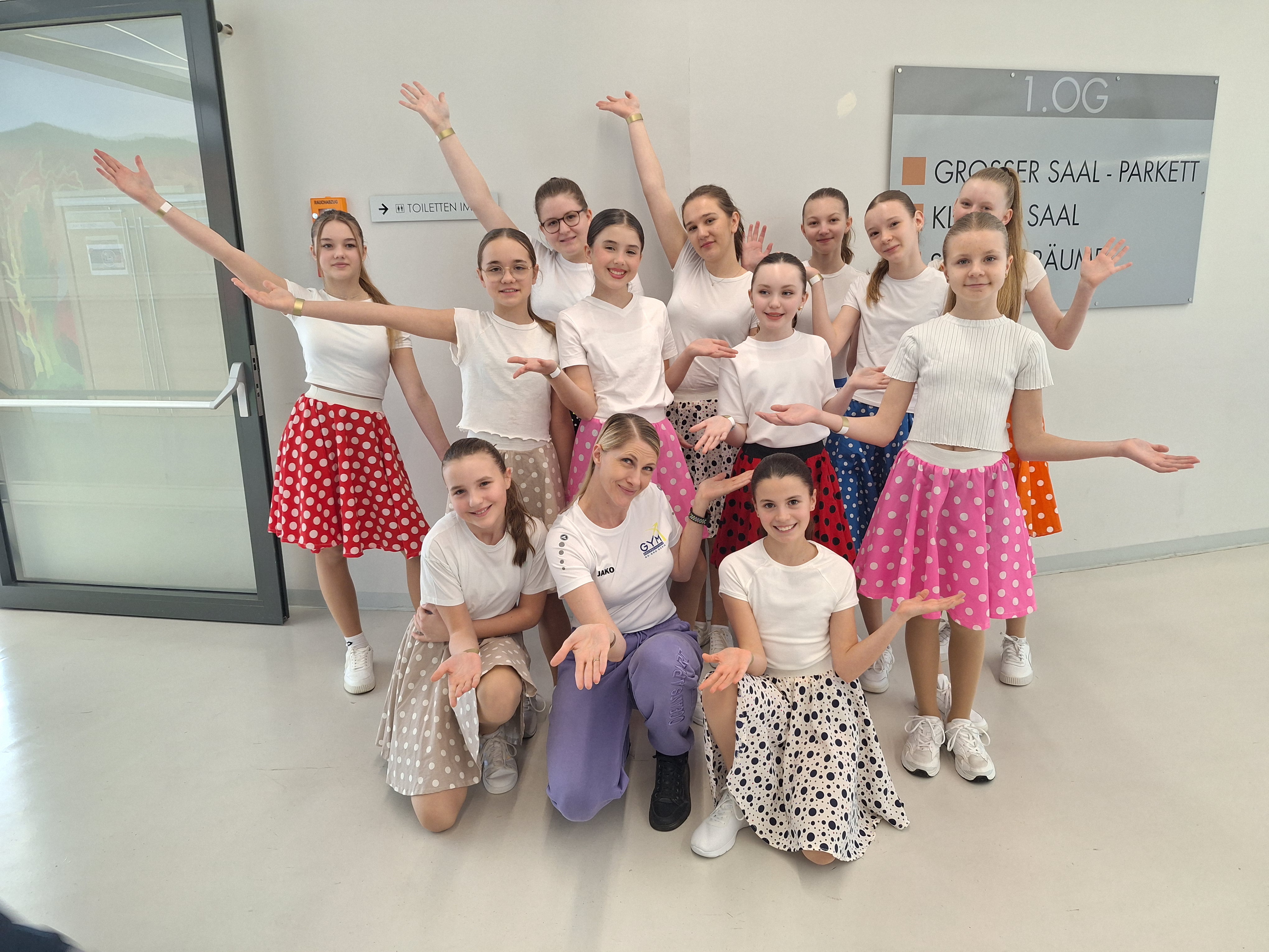 Das Bild zeigt eine Gruppe von jungen Tänzerinnen und einer erwachsenen Frau, die wahrscheinlich ihre Trainerin ist. Insgesamt sind **elf Mädchen und eine Trainerin** zu sehen. Sie stehen und knien zusammen und schauen lächelnd in die Kamera.  Die Mädchen tragen **weiße T-Shirts, bunte Röcke mit Punkten und weiße Turnschuhe**. Die Röcke sind in verschiedenen Farben, zum Beispiel **rot, rosa, blau, beige und orange**. Viele der Mädchen haben ihre **Arme nach oben oder zur Seite ausgestreckt**, als würden sie eine Tanzpose zeigen.  In der Mitte vorne kniet die **Trainerin**. Sie trägt ein **weißes T-Shirt, eine lila Hose und schwarze Schuhe**. Auch sie hält ihre Hände offen nach vorne, ähnlich wie die Mädchen. Alle wirken **fröhlich und stolz**, als hätten sie gerade einen Auftritt gehabt.  Im Hintergrund sieht man eine **helle Wand mit einem Schild**, auf dem auf Deutsch **„1. OG – Großer Saal – Parkett – Kleiner Saal“** steht. Außerdem ist links eine **Glastür** zu sehen.  Das Foto wurde wahrscheinlich **vor oder nach einer Tanzaufführung oder einem Tanzwettbewerb** aufgenommen. Die Gruppe wirkt gut organisiert und präsentiert gemeinsam ihre Tanzpose.