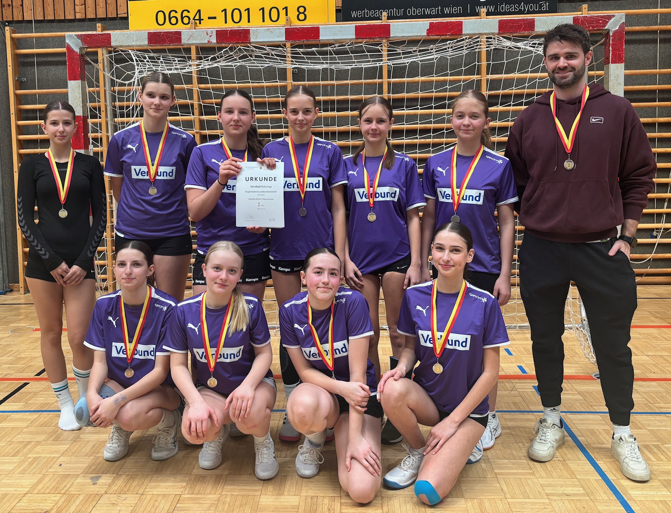 Das Foto zeigt eine Mädchen-Handballmannschaft in einer Sporthalle. Die Gruppe steht vor einem Handballtor mit Netz. Insgesamt sind zehn Spielerinnen und ein Trainer zu sehen.  Die Spielerinnen tragen einheitliche violette Trikots mit einem weißen Logo auf der Brust sowie schwarze Sporthosen. Alle haben Medaillen mit rot-gelben Bändern um den Hals. Eine Spielerin in der Mitte der hinteren Reihe hält eine Urkunde in der Hand, auf der „Urkunde“ zu lesen ist.  Fünf Spielerinnen stehen in der hinteren Reihe, während vier Spielerinnen davor knien. Der Trainer steht rechts neben der Mannschaft und trägt ebenfalls eine Medaille. Alle blicken in die Kamera und lächeln.  Im Hintergrund erkennt man den Holzboden der Sporthalle, das Handballtor sowie Werbetafeln über dem Tor. Das Foto zeigt einen stolzen Moment nach einem erfolgreichen Handballturnier.