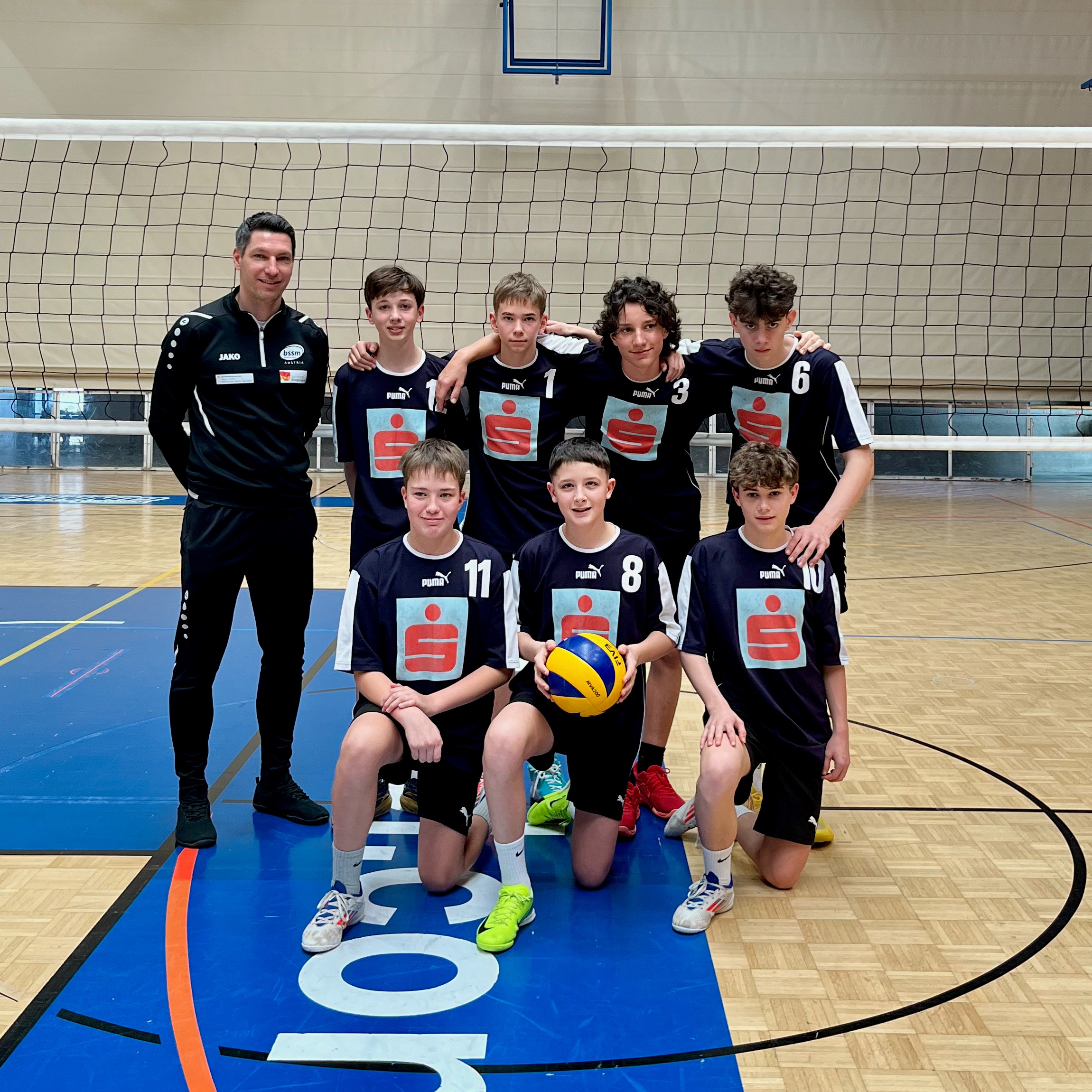 Das Bild zeigt eine Volleyballmannschaft von Jugendlichen in einer Sporthalle. Insgesamt sind acht Spieler sowie ein Trainer zu sehen. Der Trainer steht links neben der Gruppe und trägt einen schwarzen Trainingsanzug.  Die Spieler tragen einheitliche dunkle Trikots mit Nummern und einem großen Logo auf der Vorderseite. Drei von ihnen knien in der vorderen Reihe, wobei einer einen Volleyball in den Händen hält. Die übrigen fünf Spieler stehen dahinter und haben teilweise die Arme freundschaftlich umeinander gelegt.  Im Hintergrund ist ein gespanntes Volleyballnetz zu sehen, dahinter die Wand der Turnhalle mit einem Basketballkorb. Der Hallenboden ist aus Holz und mit Spielfeldmarkierungen versehen.  Die Gruppe wirkt geschlossen und motiviert, als hätten sie gerade ein Spiel oder ein Training gemeinsam absolviert.