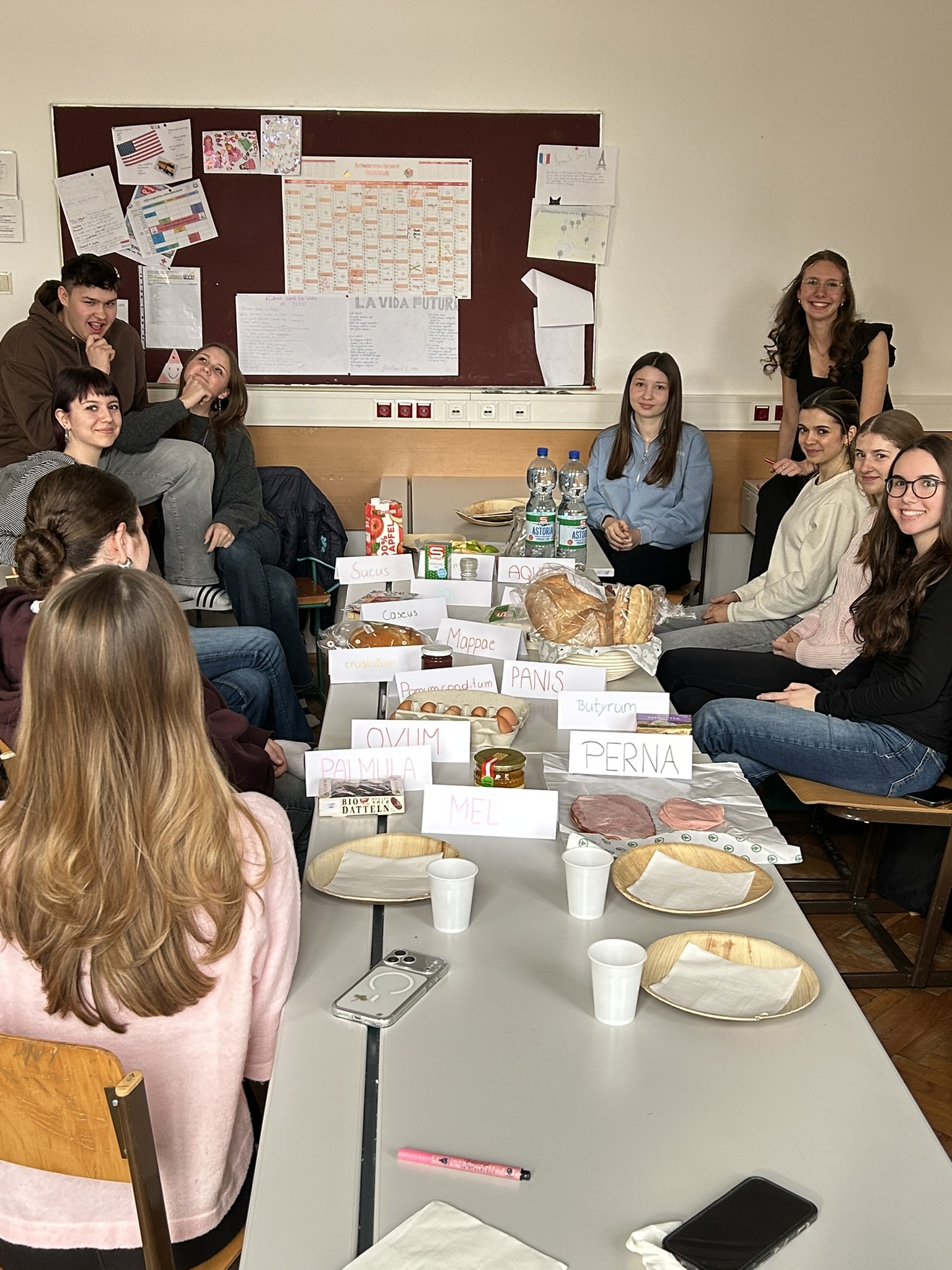 Auf dem Bild sieht man eine Gruppe von Schülerinnen und Schülern in einem Klassenraum, die gemeinsam an einem Tisch sitzen. In der Mitte stehen mehrere zusammengeschobene Tische, auf denen ein Frühstück vorbereitet ist. Darauf befinden sich verschiedene Lebensmittel wie Brot, Eier, Schinken, Butter, Käse, Datteln, Honig und Getränke. Neben den Lebensmitteln stehen kleine Schilder mit lateinischen Bezeichnungen, zum Beispiel „panis“ (Brot), „ovum“ (Ei), „mel“ (Honig), „perna“ (Schinken), „caseus“ (Käse) und „succus“ (Saft).  Um den Tisch herum sitzen mehrere Schülerinnen und Schüler auf Stühlen und Bänken. Einige schauen in die Kamera und lächeln, während andere miteinander reden oder sich gegenseitig ansehen. Die Atmosphäre wirkt entspannt und freundlich, als würden sie gemeinsam frühstücken oder eine Präsentation vorbereiten.  Im Hintergrund befindet sich eine große Pinnwand mit verschiedenen Zetteln, Zeichnungen und einem Kalender. Der Klassenraum wirkt hell und ordentlich. Auf den Tischen liegen außerdem Teller, Becher und Servietten, sodass das Frühstück gleich beginnen kann.  Insgesamt zeigt das Bild vermutlich eine schulische Aktivität oder ein Projekt, bei dem die Klasse ein Frühstück vorbereitet hat und die Lebensmittel mit lateinischen Begriffen beschriftet wurden, möglicherweise im Rahmen des Lateinunterrichts.