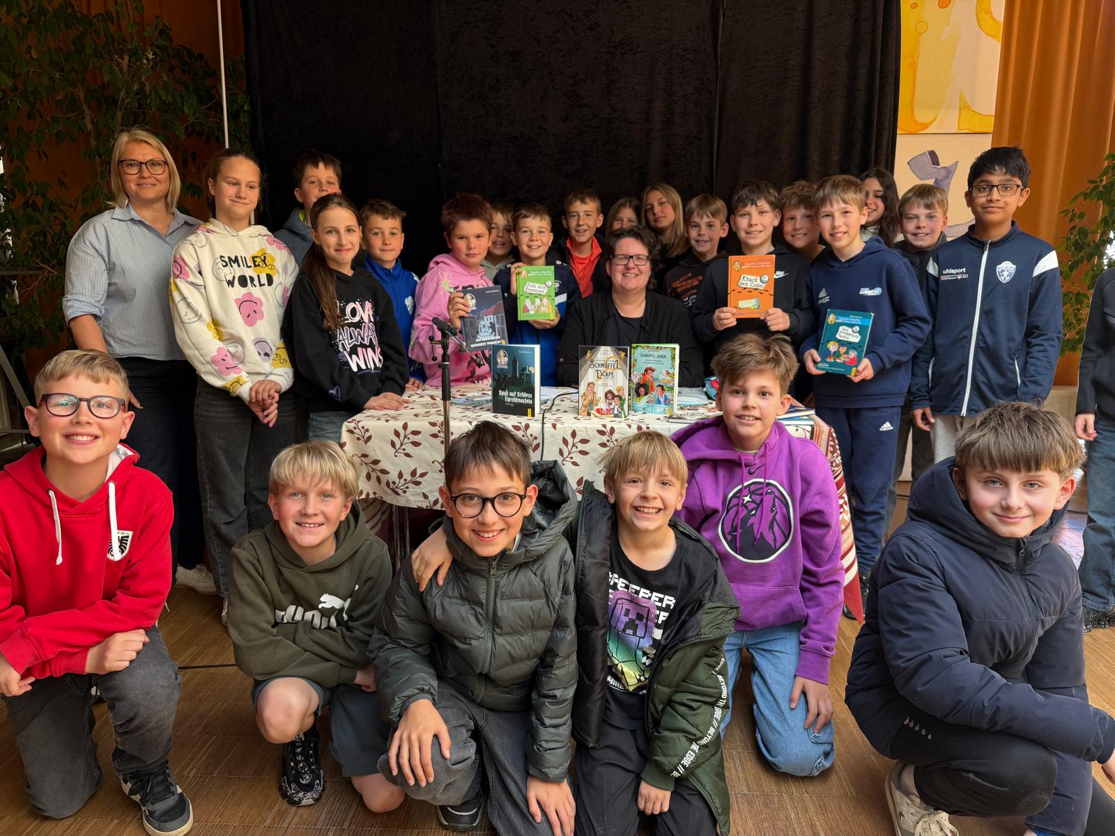 Das Bild zeigt eine große Gruppe von Schülerinnen und Schülern gemeinsam mit zwei Erwachsenen bei einer Veranstaltung in einem Raum mit Bühne. In der Mitte sitzt eine Frau an einem Tisch, auf dem mehrere Kinderbücher ausgestellt sind. Sie lächelt in die Kamera und scheint im Mittelpunkt der Veranstaltung zu stehen. Neben ihr stehen und knien zahlreiche Kinder, die ebenfalls fröhlich in die Kamera schauen. Einige von ihnen halten Bücher in den Händen und präsentieren sie stolz.  Die Kinder sind unterschiedlich gekleidet, viele tragen bunte Pullover oder Jacken, was dem Bild eine lebendige und freundliche Atmosphäre verleiht. Im Vordergrund knien einige Schüler, während die anderen hinter dem Tisch und daneben stehen. Im Hintergrund ist ein dunkler Vorhang zu sehen, der auf eine Bühne oder einen Veranstaltungsraum hinweist.  Insgesamt vermittelt das Bild den Eindruck eines gelungenen gemeinsamen Events, bei dem Lesen, Bücher und Begegnung im Mittelpunkt standen.