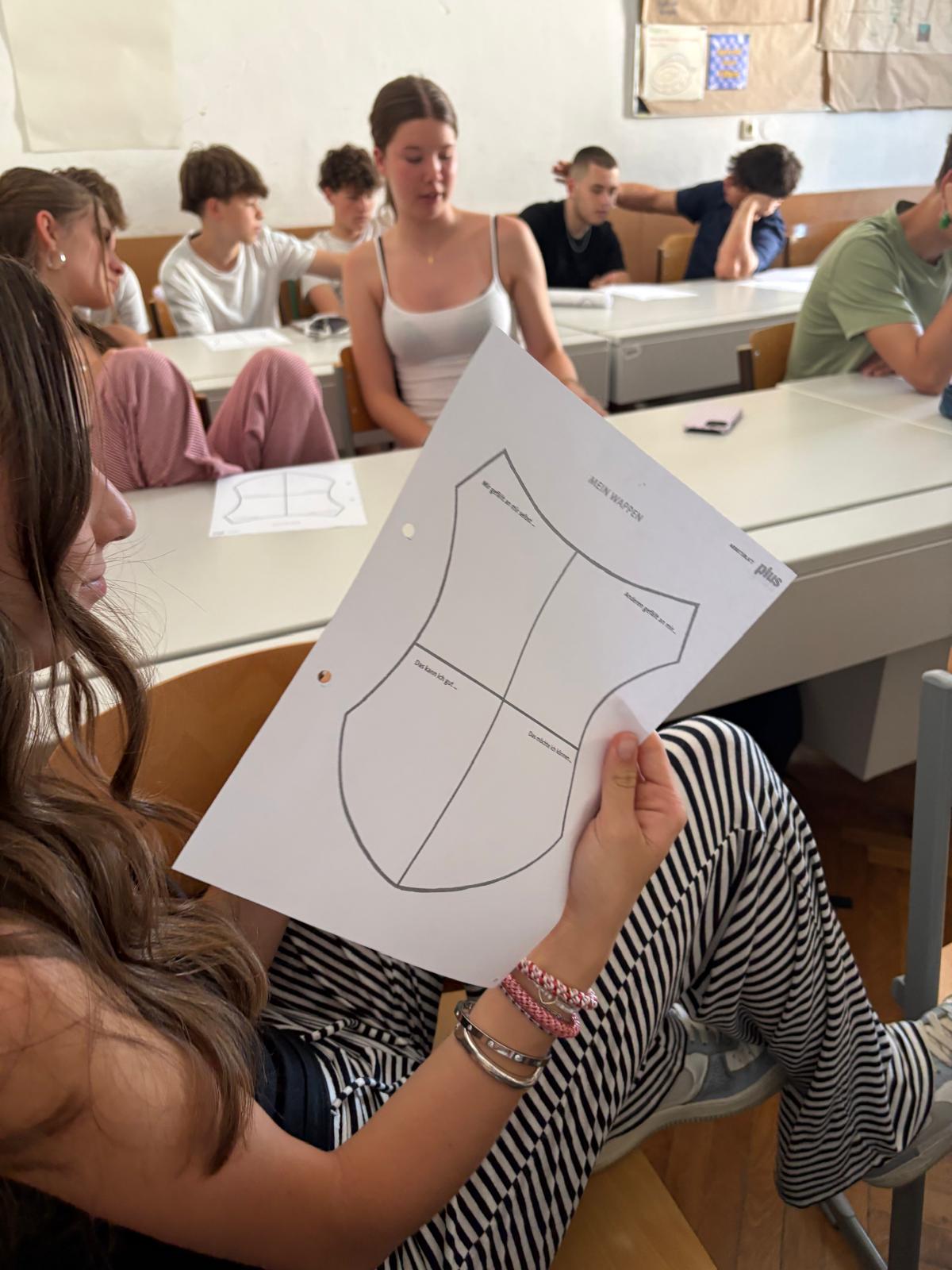 Auf dem Bild ist eine Gruppe von Jugendlichen in einem Klassenraum während eines Workshops oder Unterrichts zu sehen. Im Vordergrund hält eine Schülerin ein Arbeitsblatt in der Hand, auf dem eine schildförmige Vorlage abgebildet ist. Mehrere Schülerinnen und Schüler sitzen an Tischen, arbeiten mit Unterlagen und sind miteinander im Gespräch. Die Atmosphäre wirkt konzentriert, zugleich aber auch locker und gemeinschaftlich. Das Foto vermittelt den Eindruck einer interaktiven Unterrichtseinheit, bei der die Jugendlichen aktiv eingebunden sind.