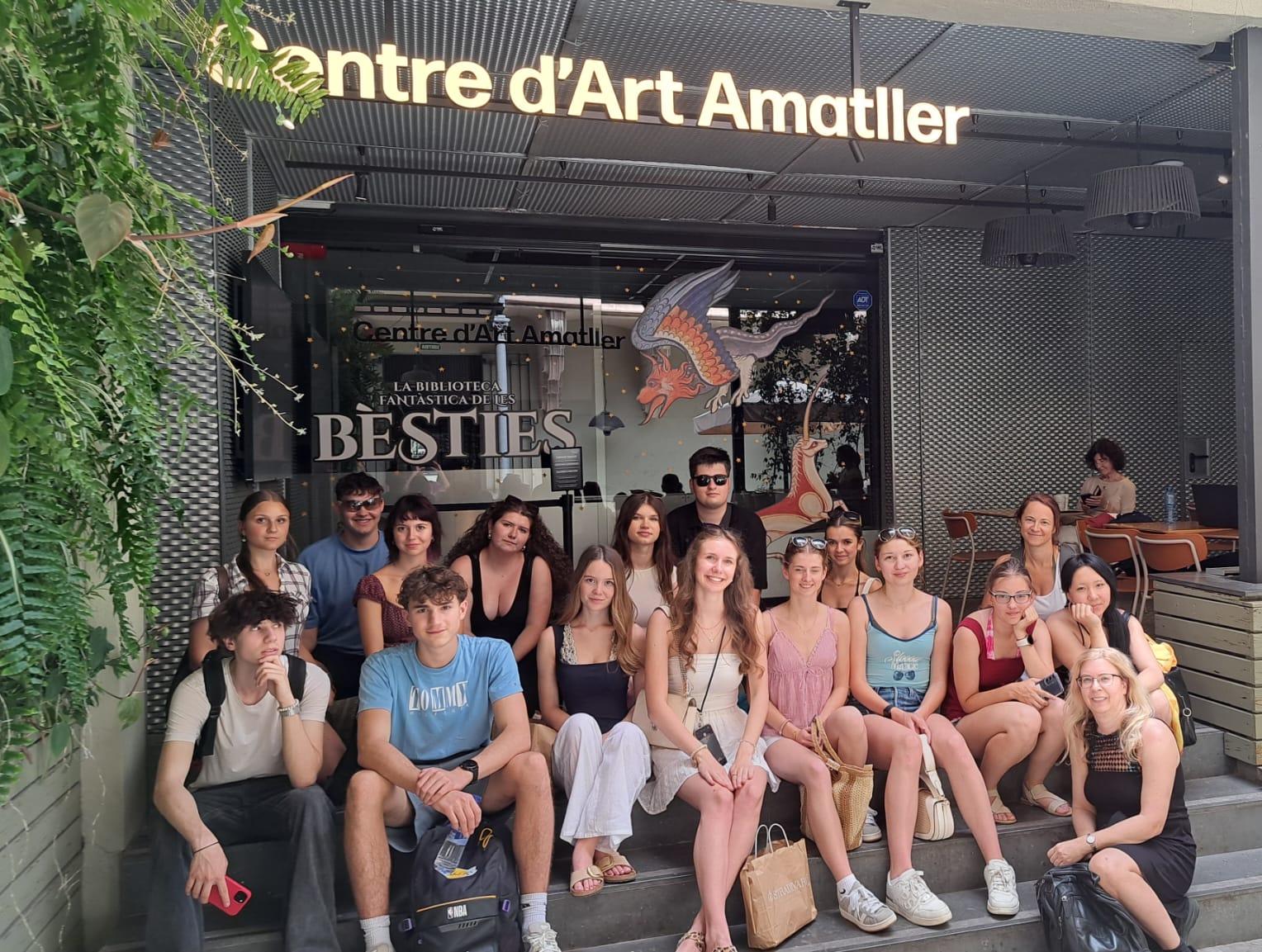 Auf dem Bild ist eine Gruppe von etwa 17 Jugendlichen und zwei erwachsenen Begleitpersonen vor dem Eingang des Centre d'Art Amatller zu sehen. Die Gruppe sitzt und steht auf den Stufen vor dem Gebäude und blickt größtenteils lächelnd in die Kamera. Die Jugendlichen tragen sommerliche Kleidung wie T-Shirts, Tops, Kleider, Shorts und Sneaker; einige haben Sonnenbrillen auf oder halten Taschen und Rucksäcke in den Händen.  Über dem Eingang leuchtet der Schriftzug „Centre d'Art Amatller“. Hinter der Glasfront ist ein großes Plakat mit der Aufschrift „La Biblioteca Fantàstica de les Bèsties“ sowie die Illustration eines fantasievollen, geflügelten Wesens zu erkennen. Links im Bild sorgen üppige grüne Pflanzen für eine natürliche Atmosphäre, während sich rechts ein überdachter Sitzbereich mit Tischen und Stühlen befindet, an dem einige Personen sitzen.  Insgesamt vermittelt das Foto den Eindruck eines gemeinsamen Ausflugs oder einer Exkursion zu einer kulturellen Einrichtung. Die Stimmung wirkt freundlich, entspannt und sommerlich.