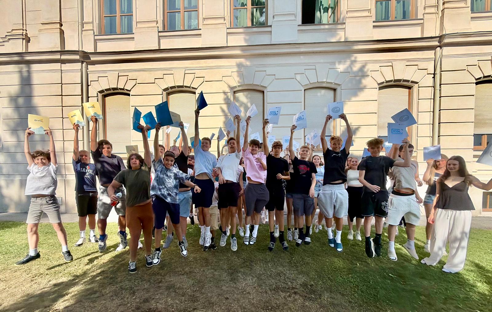 Etwa 25–30 Jugendliche sind auf einer Rasenfläche versammelt. Viele springen gleichzeitig in die Luft und halten Mappen oder Zertifikate über ihre Köpfe. Dadurch wirkt das Bild sehr lebendig und fröhlich. Die Jugendlichen tragen überwiegend sommerliche Freizeitkleidung wie T-Shirts, Shorts, Turnschuhe und Sandalen. Die Stimmung ist ausgelassen – viele lachen oder jubeln, was auf einen besonderen Anlass wie eine Abschlussfeier oder die Übergabe von Urkunden hindeutet. Im Hintergrund befindet sich ein großes, klassisches Gebäude mit einer hellen Fassade, hohen Fenstern und dekorativen Verzierungen. Das warme Sonnenlicht und die Schatten der Bäume lassen vermuten, dass das Foto an einem sonnigen Tag aufgenommen wurde.