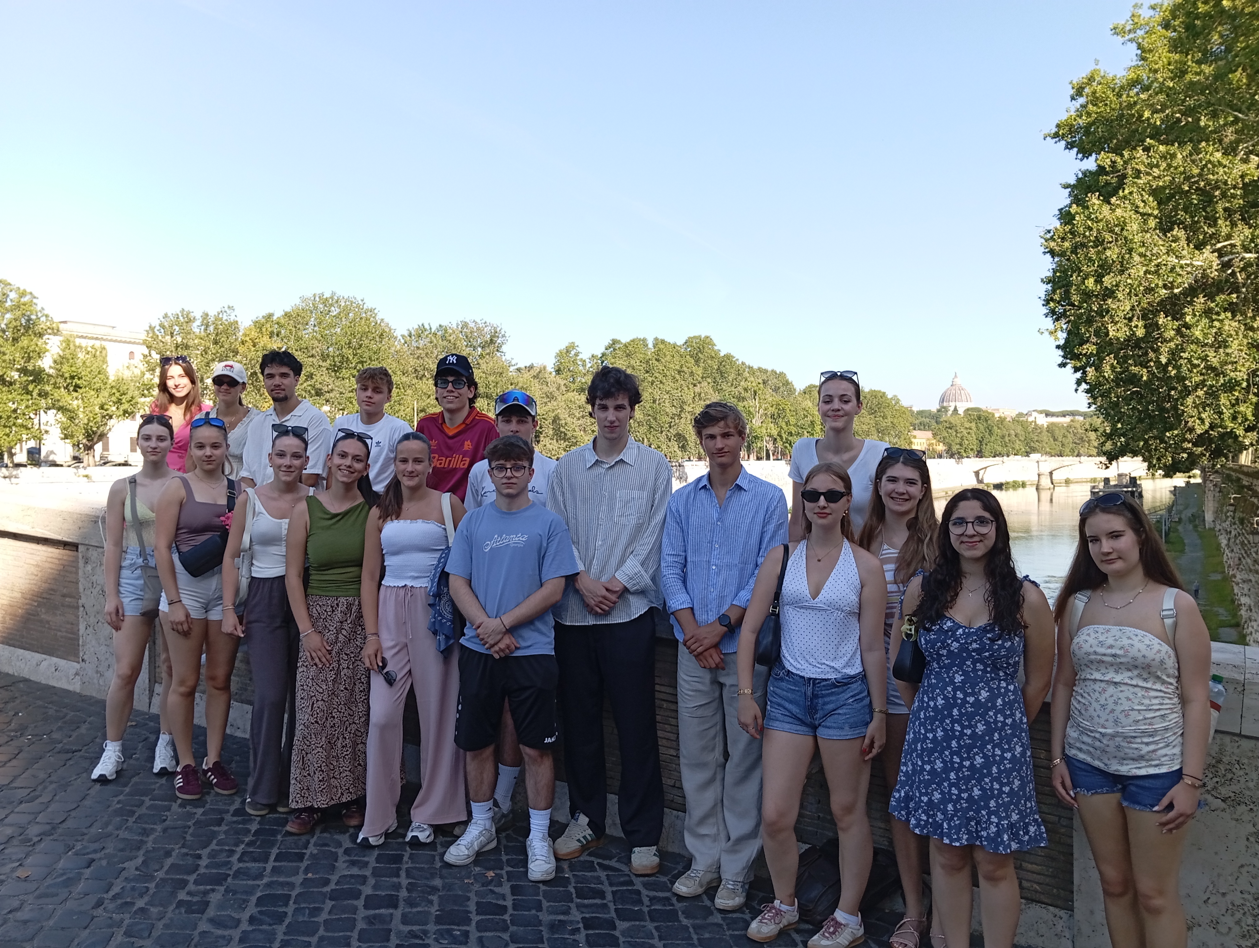 Auf dem Bild ist eine Gruppe von etwa zwanzig Jugendlichen zu sehen, die gemeinsam für ein Erinnerungsfoto posieren. Sie stehen auf einer steinernen Ufermauer an einem Fluss und blicken überwiegend lächelnd in die Kamera. Die Personen tragen sommerliche Kleidung wie T-Shirts, Blusen, Kleider, Shorts und leichte Hosen. Einige haben Sonnenbrillen oder Kappen auf. Im Hintergrund erstreckt sich eine grüne Baumreihe entlang des Flusses. Rechts ist eine Brücke zu erkennen, und in der Ferne ragt die Kuppel eines großen historischen Gebäudes über die Bäume hinaus. Der Himmel ist wolkenlos und strahlend blau, was auf einen sonnigen und warmen Tag schließen lässt. Insgesamt vermittelt das Foto den Eindruck eines gemeinsamen Ausflugs oder einer Klassenreise in einer historischen Stadt.