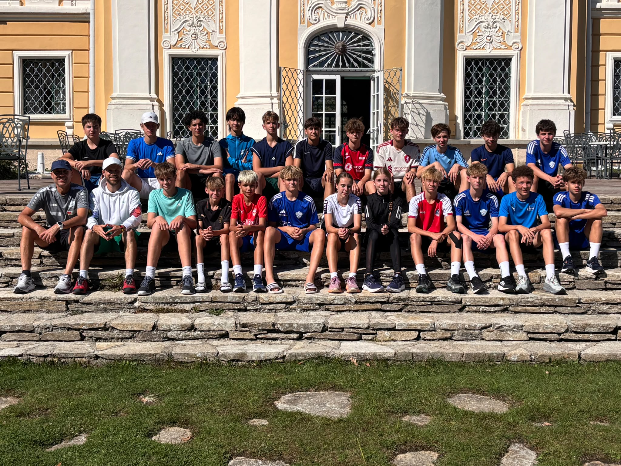 Auf dem Foto ist eine **Gruppe von Jugendlichen und zwei erwachsenen Begleitpersonen** zu sehen, die gemeinsam auf den Steinstufen vor einem historischen Gebäude sitzt bzw. steht. Die Jugendlichen tragen überwiegend **Sportkleidung und Fußballtrikots** verschiedener Vereine. Einige tragen Kappen.  Im Hintergrund ist eine **prächtig verzierte, historische Fassade** in Gelb, Weiß und Beige zu erkennen. Besonders auffällig sind die kunstvollen Stuckverzierungen, die großen Fenster mit dekorativen Gittern und eine geöffnete Tür in der Mitte.  Die Gruppe wirkt entspannt und freundlich und hat sich für ein **gemeinsames Gruppenfoto** aufgestellt. Im Vordergrund befindet sich eine Rasenfläche mit einzelnen flachen Steinen. Die Aufnahme entstand bei **hellem Sonnenschein** und vermittelt eine sommerliche, gesellige Atmosphäre.