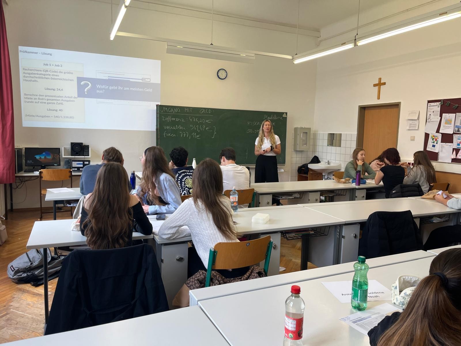 Das Bild zeigt eine Unterrichtsszene in einem Klassenzimmer. Im Vordergrund sitzen mehrere Schülerinnen und Schüler an weißen Tischen und blicken nach vorne zur Tafel. Die Lehrerin steht vorne im Raum neben einer grünen Tafel, auf der einige Begriffe und Zahlen mit Kreide geschrieben sind. An der Wand hinter ihr hängt ein Kreuz, was darauf hindeutet, dass es sich um eine Schule mit christlichem Hintergrund handeln könnte.  Links an der Wand wird eine Präsentation mit einem Beamer projiziert, auf der es um das Thema „Umgang mit Geld“ geht. Die Schülerinnen und Schüler wirken aufmerksam und beschäftigen sich mit Arbeitsmaterialien. Im Hintergrund sitzen noch weitere Schülergruppen, die miteinander sprechen oder arbeiten. Das Klassenzimmer ist hell und freundlich eingerichtet, mit großen Fenstern und moderner Beleuchtung. Auf den Tischen stehen Wasserflaschen und Arbeitsblätter, was auf eine aktive Unterrichtsatmosphäre hinweist.