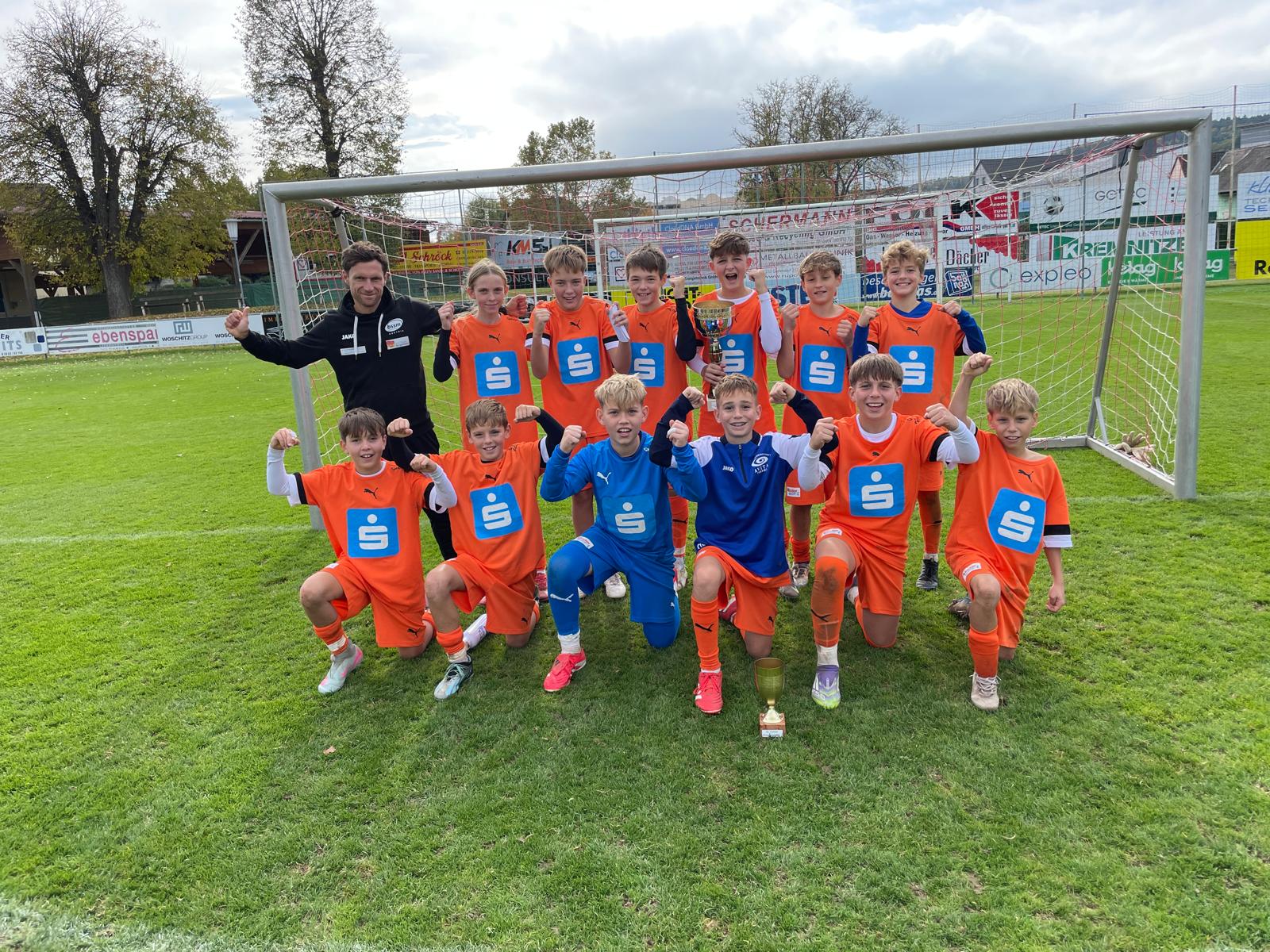 Auf dem Foto posiert eine junge Fußballmannschaft gemeinsam mit ihrem Trainer vor einem Tor auf einem grünen Rasenplatz. Die Spieler tragen leuchtend orange Trikots mit blauem Logo, zwei Jungs in der Mitte stechen in blauen Outfits hervor. Alle knien oder stehen in einer Reihe und zeigen jubelnd ihre Fäuste, vor ihnen steht ein kleiner goldener Pokal. Im Hintergrund sind Werbebanden und herbstliche Bäume zu sehen, der Himmel ist leicht bewölkt. Die Stimmung wirkt ausgelassen und stolz – ein Moment nach einem erfolgreichen Turnier oder Spiel.