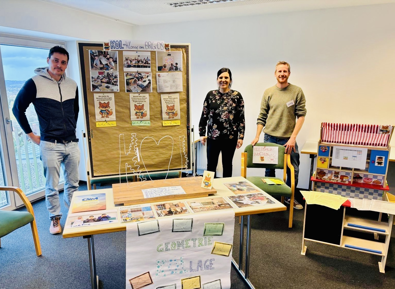 Drei Personen stehen in einem hellen Raum hinter einem Präsentationstisch. Links befindet sich eine große Pinnwand mit der Überschrift „KOL‑Klasse am BRG/OS“. Darauf sind mehrere Fotos, Plakate und kleine Fuchs‑Illustrationen angebracht. In der Mitte steht eine Frau, links und rechts von ihr jeweils ein Mann; alle lächeln in die Kamera.  Vorne steht ein Tisch mit ausgestellten Materialien: Fotos, ein DIN‑A4‑Heft, kleine Kärtchen und ein großes Plakat mit der Aufschrift „GEOMETRIE KOLLAGE“. Daneben steht ein Stuhl mit einem Schild „Feedback‑Karten“. Rechts im Bild sieht man ein weiteres Präsentationsmöbel im Stil eines kleinen Marktstandes mit rot‑weiß gestreifter Markise und zusätzlichen Plakaten. Der Raum ist freundlich, mit großem Fenster links, und vermittelt den Eindruck einer Schul‑ oder Projektpräsentation.