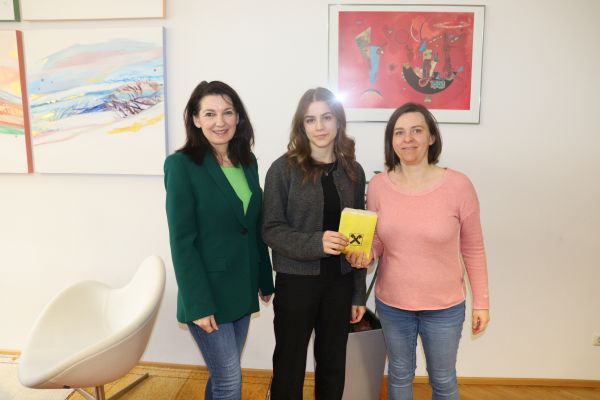 Das Bild zeigt drei Frauen, die in einem hellen Raum nebeneinander stehen und in die Kamera lächeln. Die Frau auf der linken Seite trägt einen grünen Blazer und eine blaue Jeans. Sie hat dunkles, schulterlanges Haar. Die Frau in der Mitte hat braunes, welliges Haar und trägt eine dunkle Jacke sowie eine schwarze Hose. Sie hält ein gelbes Kuvert mit dem Logo der Raiffeisenbank in den Händen, was auf eine symbolische Übergabe hinweist. Die Frau auf der rechten Seite trägt ein rosafarbenes Oberteil und eine blaue Jeans.   Im Hintergrund sind bunte Gemälde an der Wand zu sehen, die den Raum lebendig und freundlich wirken lassen. Auf der linken Seite steht ein weißer moderner Stuhl, der Teil der Einrichtung ist. Die Szene vermittelt eine positive und offizielle Atmosphäre.
