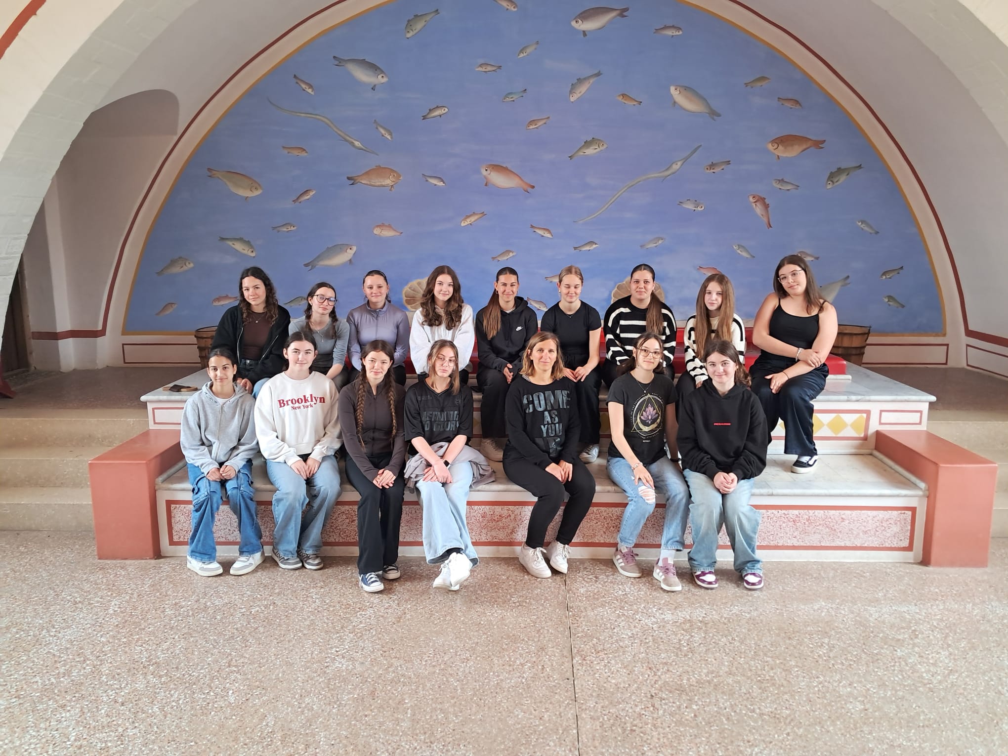 Das Bild zeigt eine Gruppe von etwa 16 jungen Frauen, die für ein Gruppenfoto in einem beeindruckenden historischen Gebäude posieren. Sie sitzen und stehen auf einer mehrstufigen Steinbank vor einer spektakulären Wandmalerei mit antikem römischen Motiv.  Die Wandmalerei zeigt einen blauen Himmel mit zahlreichen realistisch gemalten Fischen, die durch die Luft zu schwimmen scheinen - ein surreales und künstlerisches Element. Die Wand ist von einem eleganten Bogen gerahmt, der mit roten und goldenen Verzierungen geschmückt ist. Die Architektur wirkt römisch-antik mit ihren klassischen Proportionen und Farben.  Die jungen Frauen tragen lässige, moderne Kleidung - hauptsächlich Jeans, Hoodies und Sneaker in verschiedenen Farben. Einige haben Aufschriften auf ihren Oberteilen wie &quot;Brooklyn&quot; und andere Motive. Sie wirken entspannt und fröhlich, als wären sie auf einem Schulausflug oder einer Bildungsreise.  Der Boden besteht aus hellem Stein oder Beton, was den antiken Charakter des Ortes unterstreicht. Basierend auf dem Dateinamen &quot;Carnuntum&quot; handelt es sich wahrscheinlich um das römische Freilichtmuseum Carnuntum in Österreich, das für seine rekonstruierten römischen Gebäude und Wandmalereien bekannt ist.