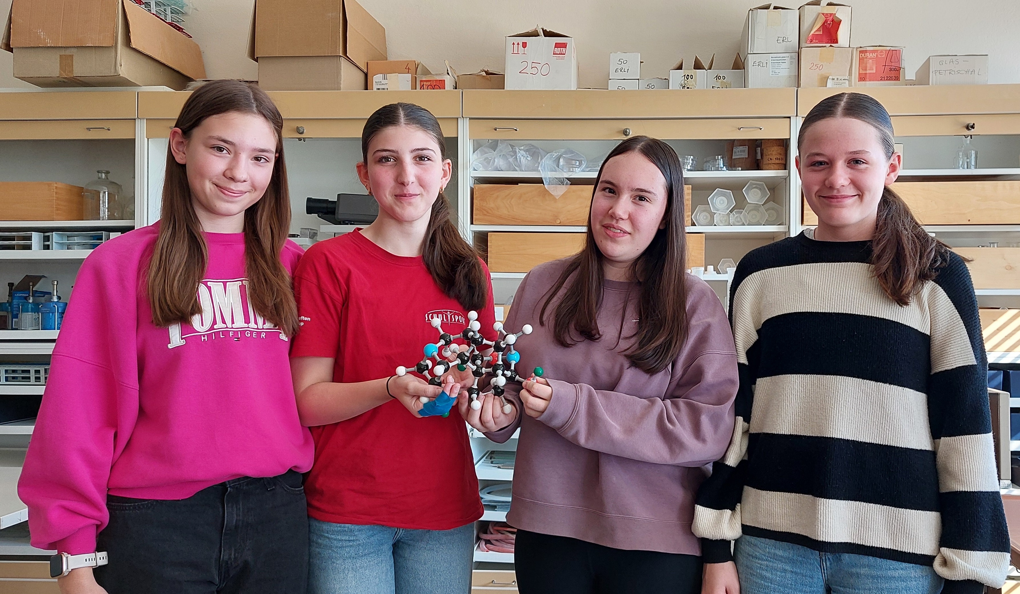 Das Bild zeigt vier Schülerinnen in einem Chemielabor, die gemeinsam ein dreidimensionales Molekülmodell präsentieren. Die Mädchen stehen nebeneinander und halten das bunte Modell aus verschiedenfarbigen Kugeln (weiß, schwarz, rot, blau, grün) und Verbindungsstäben in den Händen.  Sie tragen unterschiedliche Kleidung: von links nach rechts ein pinkes &quot;JMU Hilfiger&quot; Sweatshirt, ein rotes Oberteil, einen rosa Hoodie und einen gestreiften Pullover in Beige-Schwarz. Alle lächeln freundlich in die Kamera.  Im Hintergrund ist die typische Ausstattung eines Schullabors zu sehen: Regale mit Chemikalienflaschen, Laborgeräten und Aufbewahrungsboxen. Die Regale sind mit nummerierten Kartons und verschiedenen Laborutensilien gefüllt, was auf einen gut ausgestatteten Chemieunterricht hindeutet.  Die Atmosphäre wirkt entspannt und lernfreudig - offensichtlich arbeiten die Schülerinnen an einem Chemieprojekt zur Molekularstruktur.