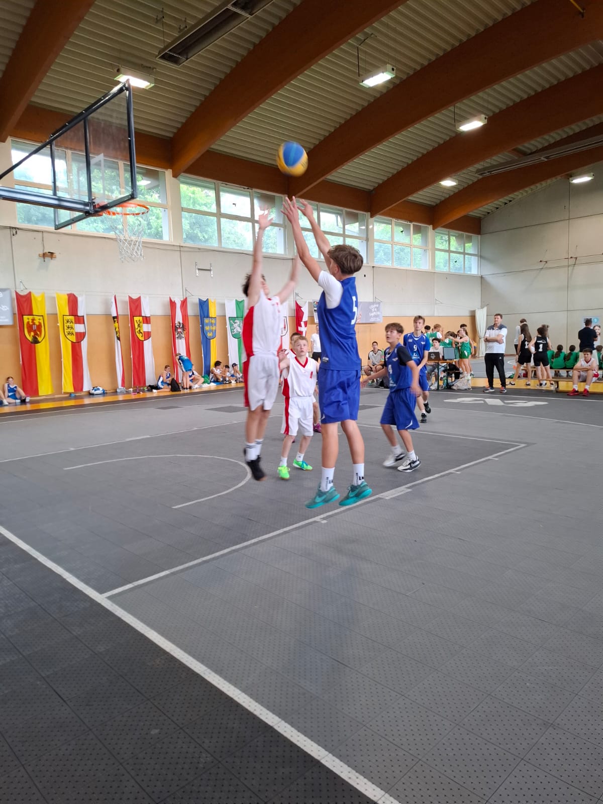 Das Bild zeigt eine lebendige Basketballszene in einer modernen Sporthalle. Im Zentrum des Geschehens springen zwei Jugendliche - einer in weißem Trikot mit roten Akzenten, der andere in blauem Trikot - gleichzeitig hoch, um einen blau-gelben Basketball zu erreichen, der sich in der Luft befindet.  Die Sporthalle beeindruckt durch ihre Architektur mit sichtbaren Holzbalken an der Decke und großen Fenstern, die natürliches Licht hereinlassen. An der Wand sind bunte Fahnen verschiedener österreichischer Bundesländer aufgehängt, was auf ein regionales Turnier hindeutet.  Auf dem grauen Hallenboden sind die weißen Linien des Basketballfeldes klar erkennbar. Im Hintergrund sitzen Zuschauer auf Bänken und verfolgen das Spiel aufmerksam. Links im Bild ist der Basketballkorb mit seinem charakteristischen orangefarbenen Ring sichtbar.  Die Atmosphäre wirkt sportlich und jugendlich - es scheint sich um ein Nachwuchsturnier oder Schulwettkampf zu handeln, bei dem junge Basketballtalente ihr Können unter Beweis stellen.