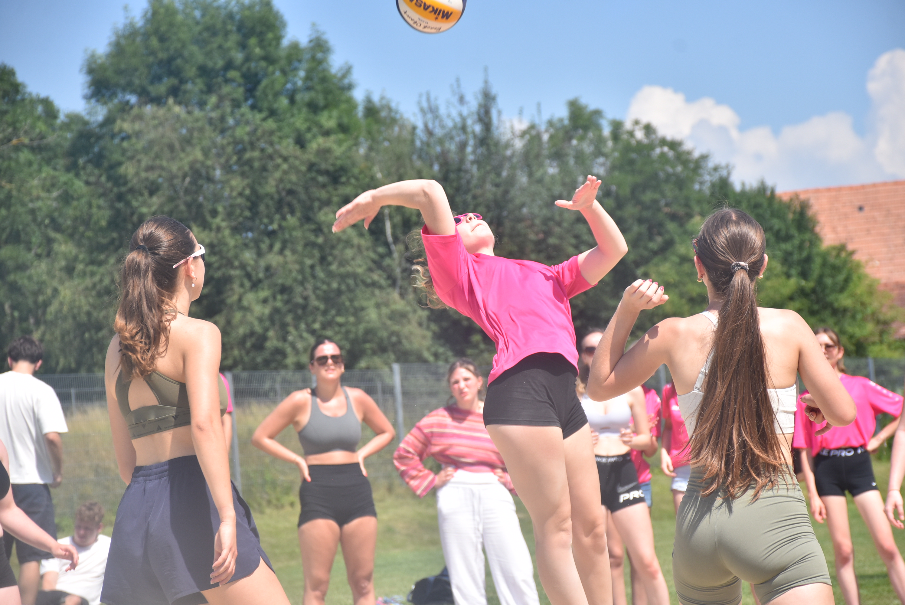 Auf dem Bild ist eine Volleyballszene im Freien zu sehen. Im Mittelpunkt springt eine Spielerin im pinken Shirt nach oben, um den Ball zu spielen. Rund um sie stehen weitere Schülerinnen auf dem Spielfeld und verfolgen aufmerksam den Ballwechsel. Im Hintergrund sind außerdem Mitschülerinnen und Mitschüler zu sehen, die das Spiel beobachten. Das sonnige Wetter, die sportliche Dynamik und die konzentrierte Stimmung vermitteln den Eindruck eines spannenden und gelungenen Volleyballmatches.