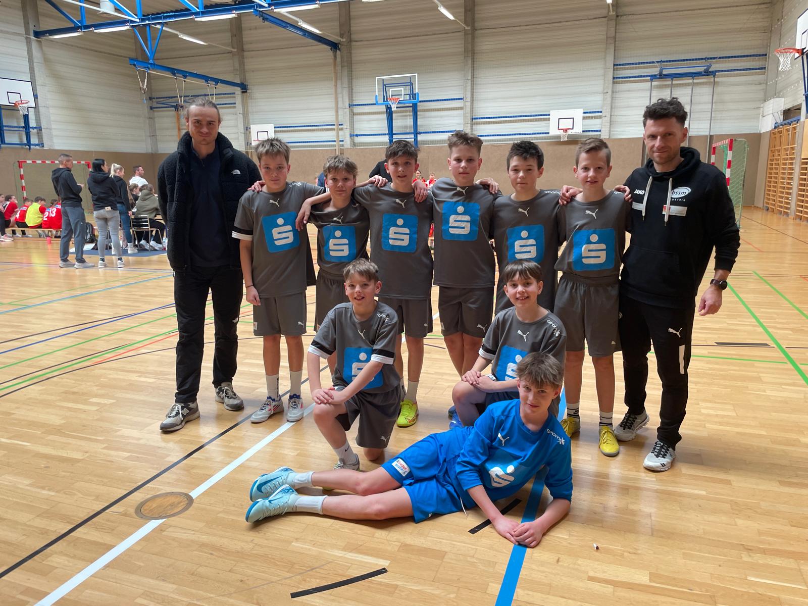 Das Bild zeigt eine Jugend-Handballmannschaft in einer Sporthalle. Die Mannschaft besteht aus neun jungen Spielern, die graue Trikots mit blauen Sparkassen-Logos auf der Brust tragen. Ein Spieler liegt im Vordergrund in einem blauen Torwarttrikot.  Die Mannschaft wird von zwei Trainern oder Betreuern in schwarzen Jacken flankiert, die links und rechts neben der Gruppe stehen. Die Aufstellung ist für ein Teamfoto arrangiert - sechs Spieler stehen in einer Reihe, zwei knien davor, und der Torwart liegt im Vordergrund.  Im Hintergrund der modernen Sporthalle sind Basketballkörbe, Handballtore mit rot-weißen Markierungen und Zuschauer oder andere Mannschaften zu erkennen. Der Hallenboden ist aus Holz und mit verschiedenfarbigen Linien für unterschiedliche Sportarten markiert.