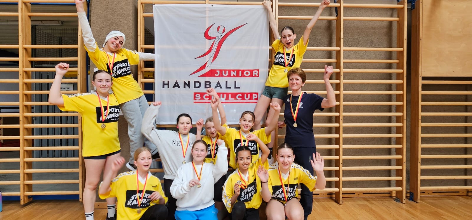 Das Bild zeigt eine jubelnde Mädchen-Handballmannschaft, die an einem &quot;Junior Handball Schulcup&quot; teilgenommen hat. Die Spielerinnen tragen überwiegend gelbe Trikots mit der Aufschrift &quot;Sport Kainrath&quot; und haben Medaillen um den Hals.  Die Mannschaft posiert vor einer Sprossenwand in einer Turnhalle, an der ein weißes Banner mit dem Logo des &quot;Junior Handball Schulcup&quot; befestigt ist. Die Mädchen zeigen verschiedene Jubelposen - einige heben die Arme hoch, andere lächeln freudig in die Kamera. Einige Spielerinnen tragen weiße Pullover statt der gelben Teamtrikots.  Rechts im Bild steht eine Trainerin oder Betreuerin in einem dunklen Shirt, die ebenfalls eine Medaille trägt und mit der Mannschaft feiert. Die Atmosphäre strahlt Teamgeist und Freude über einen gemeinsamen Erfolg aus.