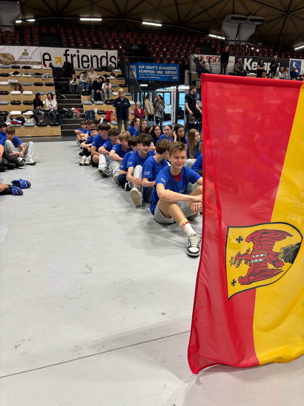 Das Bild zeigt eine Sportveranstaltung in einer Sporthalle oder Arena. Im Vordergrund sitzen junge Sportler in blauen Trikots in einer Reihe auf dem Boden. Rechts im Bild ist eine deutsche Flagge mit dem Bundesadler zu sehen. Im Hintergrund befinden sich Zuschauerränge mit einigen Besuchern und Werbebannern, darunter ein &quot;e-friends&quot; Banner und ein deutsches Motivationsbanner mit dem Text &quot;Auf geht's Burschen, kämpfen und siegen!&quot;. Die Atmosphäre wirkt wie bei einem Schulwettkampf oder einer Jugend-Sportveranstaltung, bei der die Teilnehmer auf ihren Einsatz warten.