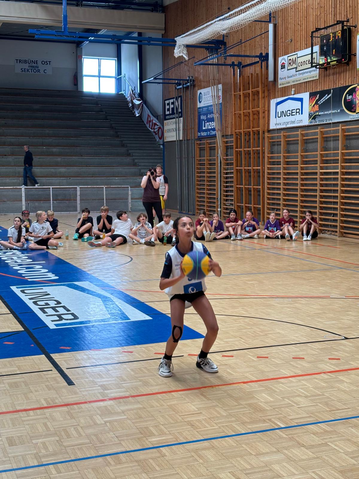 Das Bild zeigt eine Sportszene in einer Sporthalle. Im Vordergrund ist ein junger Spieler in einem blau-weißen Trikot zu sehen, der einen gelb-blauen Volleyball hält und sich auf das Spiel konzentriert. Der Spieler trägt eine Kniebandage am rechten Knie und Sportschuhe.  Im Hintergrund sitzen mehrere Kinder und Jugendliche auf dem Hallenboden an der Wand und beobachten das Geschehen. Die Sporthalle hat einen typischen Parkettboden mit farbigen Markierungen für verschiedene Sportarten. An den Wänden sind Sponsorenbanner zu sehen, darunter &quot;UNGER&quot; und andere Firmenlogos.  Links im Bild ist eine Tribüne mit Stufen erkennbar, auf der &quot;TRIBÜNE OST SEKTOR C&quot; steht. Die Halle ist mit Sprossenwänden und einem Volleyballnetz ausgestattet. Eine Person mit Kamera steht am Rand und dokumentiert das Ereignis. Die gesamte Szene wirkt wie ein Jugend-Volleyballturnier oder -training.