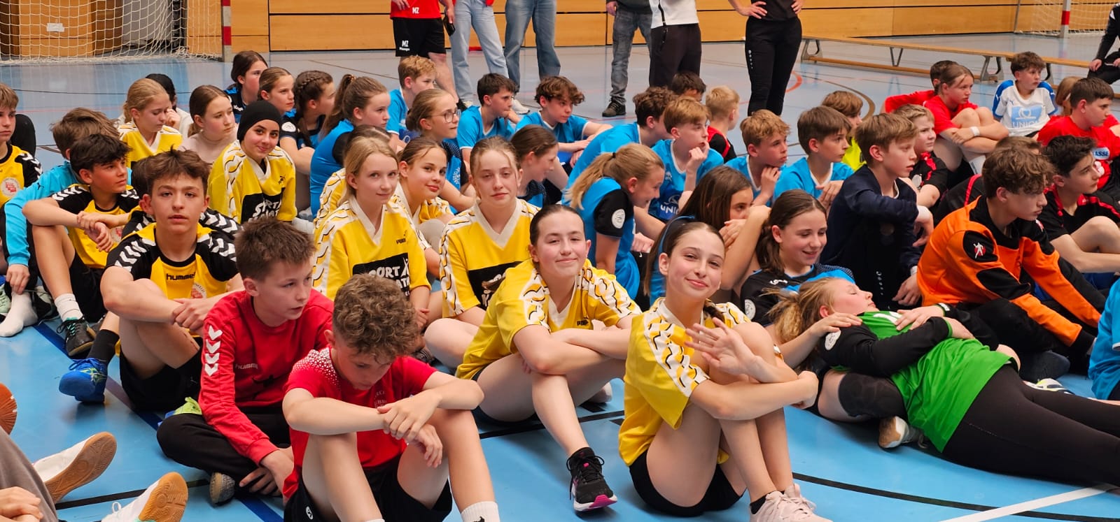 Das Bild zeigt eine Handballhalle mit jungen Spielerinnen und Spielern, die auf dem Hallenboden sitzen. Im Vordergrund sind mehrere Jugendliche in verschiedenfarbigen Trikots zu sehen - einige in gelben, andere in roten und blauen Trikots. Sie sitzen in Gruppen auf dem blauen Hallenboden vor einem Handballtor mit rot-weißer Markierung. Im Hintergrund ist das Handballtor mit seinem Netz deutlich erkennbar, und über dem Spielfeld befinden sich einige Zuschauer auf einer erhöhten Tribüne. Die Szene wirkt wie ein Jugendturnier oder eine Trainingsveranstaltung, bei der die Teams eine Pause machen oder auf ihren Einsatz warten.