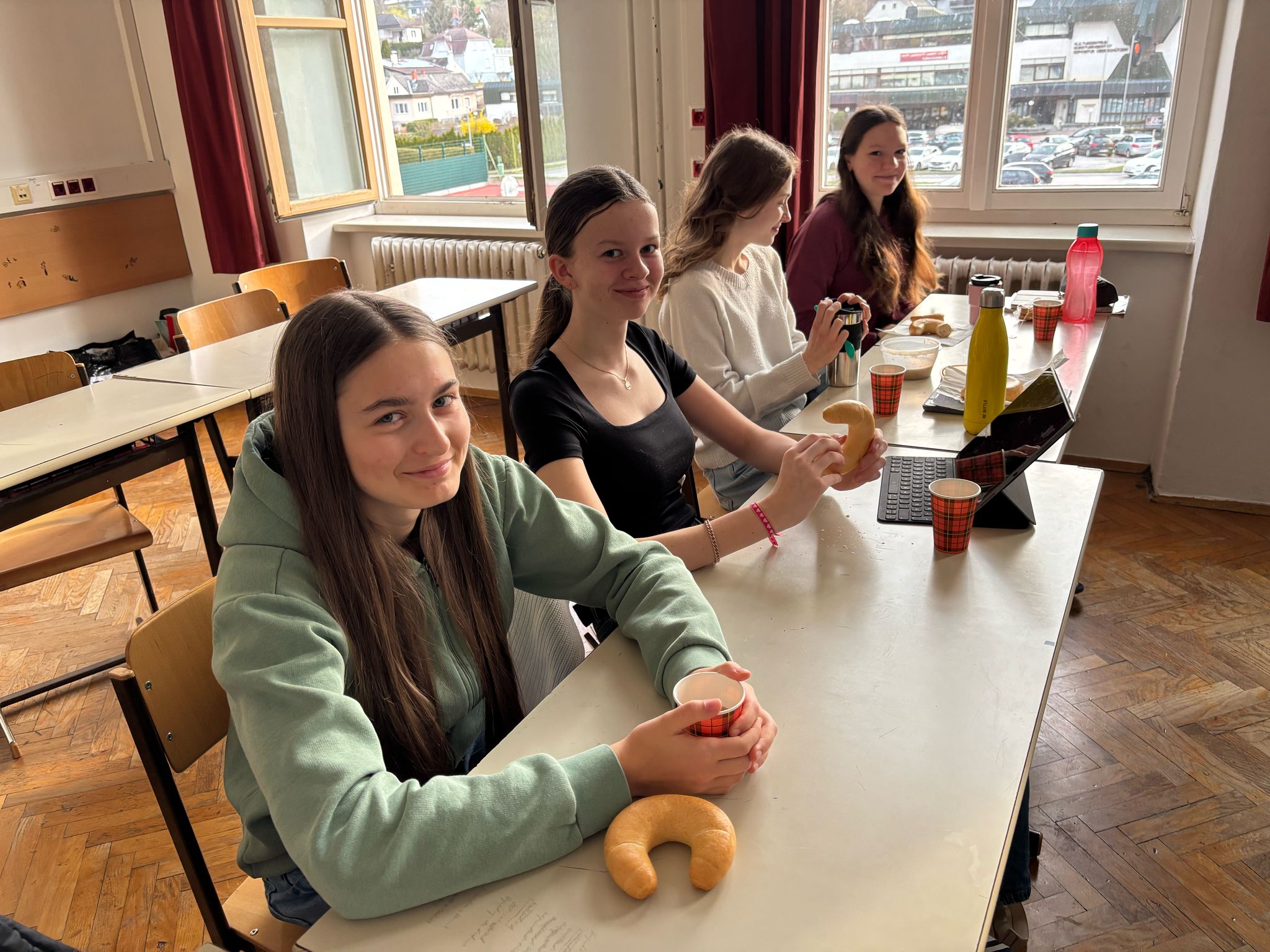 Das Bild zeigt einen Klassenraum mit vier jungen Personen, die an Schultischen sitzen. Im Vordergrund sitzt eine Person in einem mintgrünen Pullover mit einem Croissant und einem Pappbecher. Dahinter sitzen drei weitere Personen - eine in einem schwarzen Oberteil, eine in einem weißen Pullover und eine in einem weinroten Oberteil.  Auf den Tischen befinden sich verschiedene Getränke in bunten Bechern und Flaschen (gelb und rot), ein Laptop und Snacks. Der Raum hat große Fenster mit roten Vorhängen, durch die man Gebäude und parkende Autos sehen kann. Der Boden besteht aus Parkettholz im Fischgrätmuster.  Die Atmosphäre wirkt entspannt, wie bei einer Pause oder einer informellen Lerngruppe.