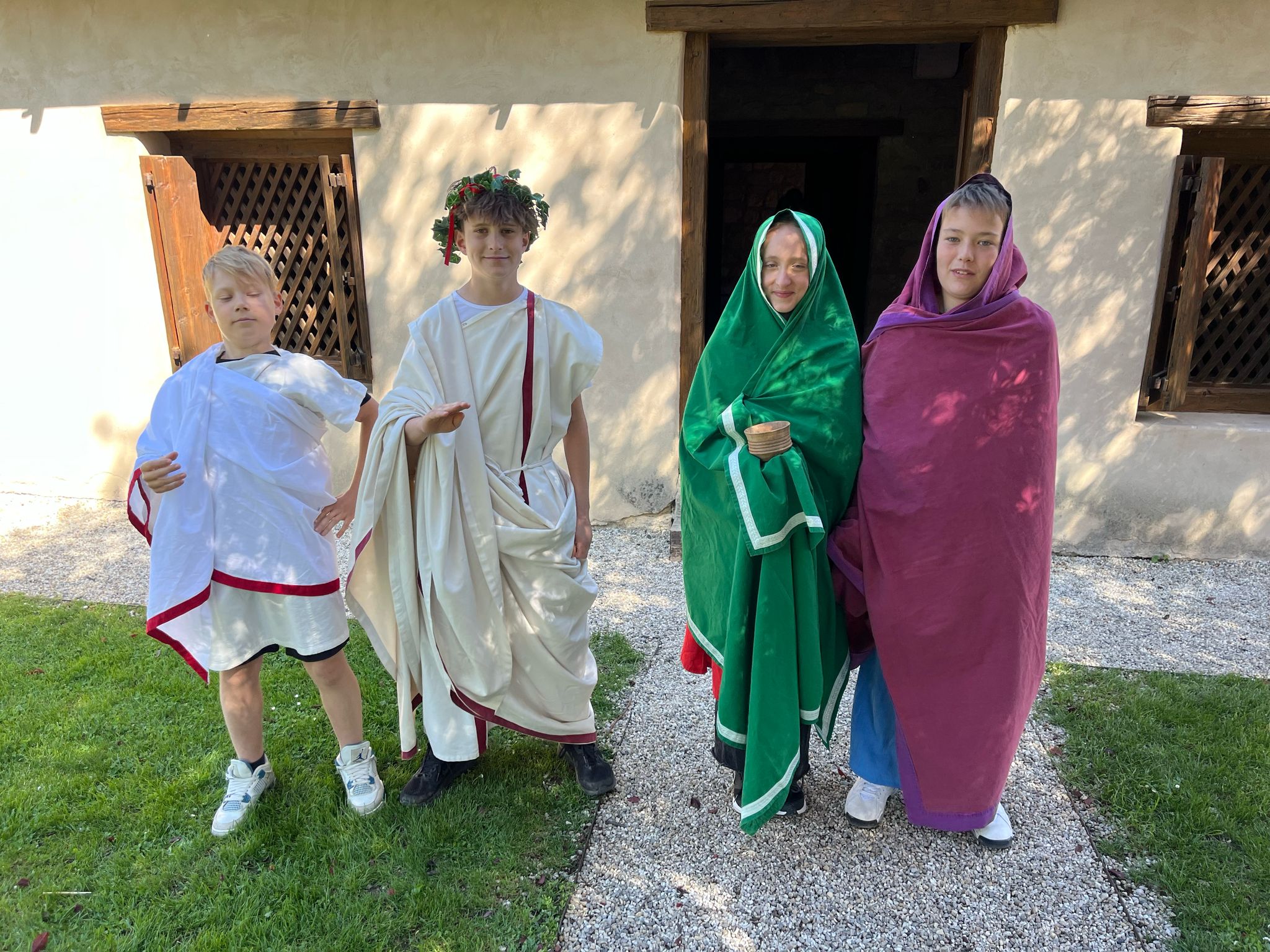 Auf dem Bild sind vier Kinder zu sehen, die in historischen römischen Gewändern verkleidet sind. Sie stehen vor einem Gebäude mit hellen Wänden, Holztüren und Fenstern mit Holzgittern. Die beiden Kinder links tragen weiße Tuniken mit roten Borten, wobei das zweite Kind von links zusätzlich einen Lorbeerkranz auf dem Kopf trägt. Die beiden Kinder rechts sind in lange Umhänge gehüllt, einer in grün mit weißem Rand, der andere in lila. Das Kind im grünen Umhang hält einen kleinen Holzbecher in der Hand. Die Kinder stehen auf einem Kiesweg, der von grünem Rasen umgeben ist, und schauen in die Kamera. Die Szene wirkt wie eine Nachstellung aus der römischen Antike, vermutlich im Rahmen eines Projekttages oder einer Schulveranstaltung.