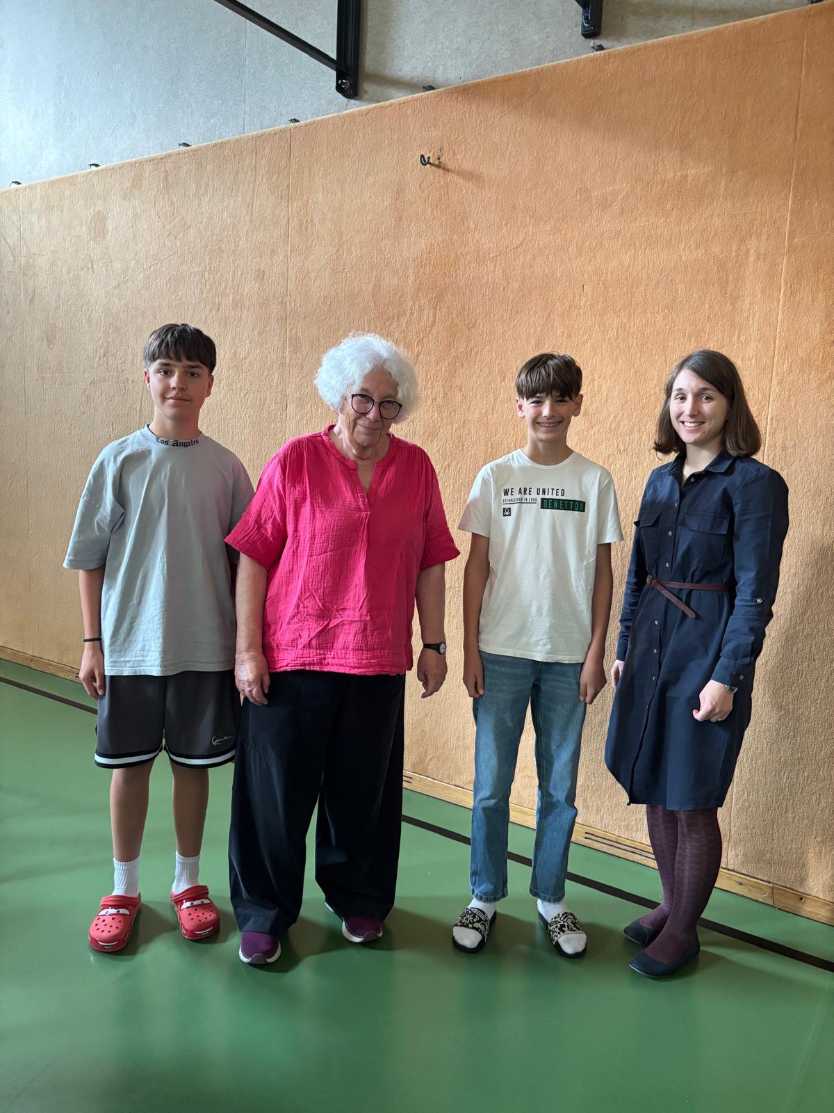 Das Bild zeigt vier Personen, die in einer Turnhalle oder einem Mehrzweckraum vor einer beigen Wand stehen. Von links nach rechts sind zu sehen: ein Jugendlicher in grauem T-Shirt und schwarzen Nike-Shorts mit roten Crocs, eine ältere Dame mit weißen lockigen Haaren, Brille und einer leuchtend pinken Bluse, ein weiterer Jugendlicher in hellem T-Shirt mit Aufdruck und Jeans, sowie eine junge Frau in einem dunkelblauen Hemdblusenkleid mit braunen Stiefeln.  Die ältere Dame steht im Zentrum der Gruppe und scheint die Hauptperson zu sein - vermutlich die im Dateinamen erwähnte &quot;Zeitzeugin&quot;. Der grüne Hallenboden und die schlichte Ausstattung deuten auf eine Schulveranstaltung oder ein Bildungsprojekt hin, bei dem die Zeitzeugin ihre Erfahrungen mit den Jugendlichen und der Betreuerin teilt.
