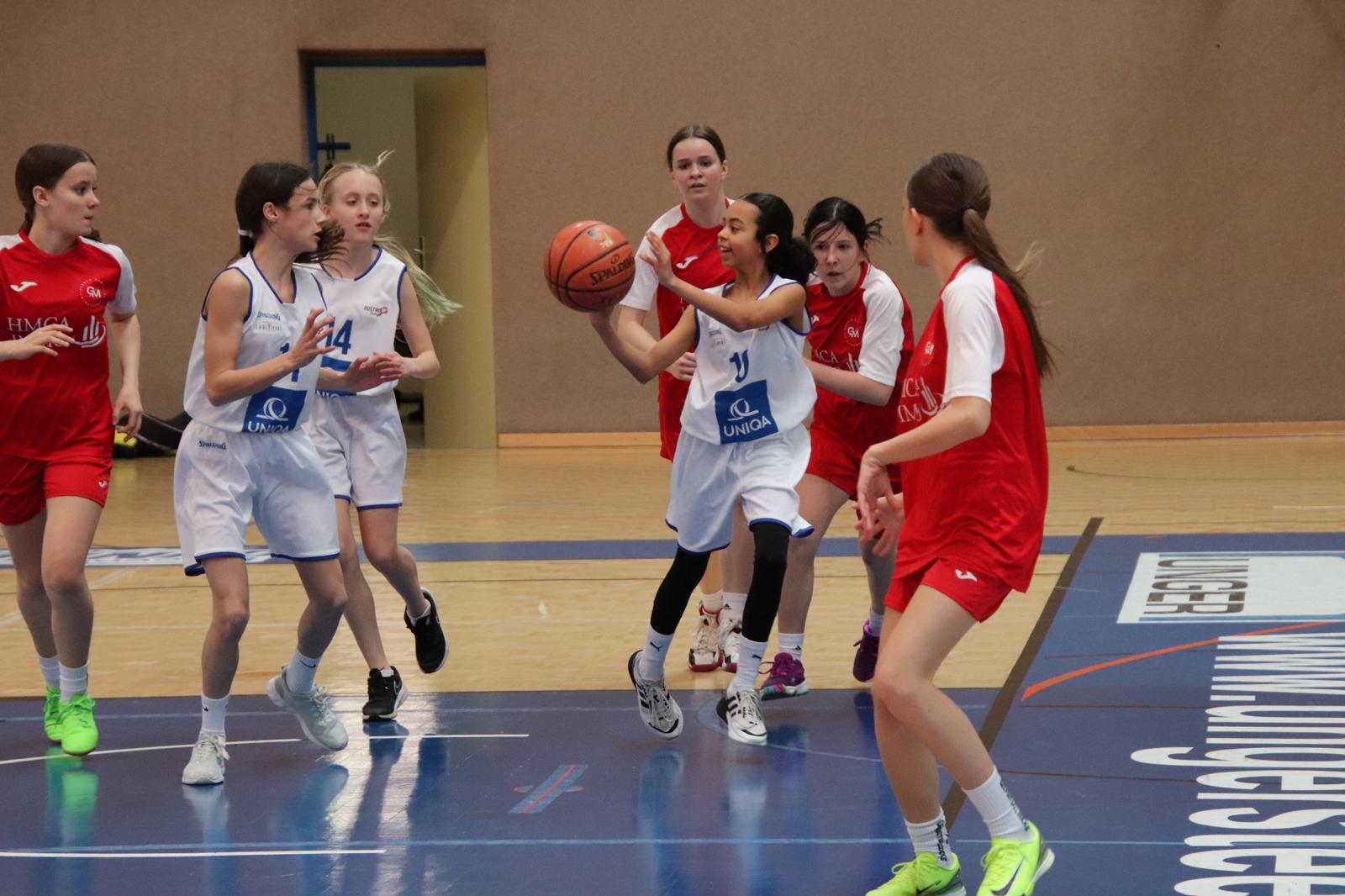 Das Bild zeigt ein Basketballspiel in einer Sporthalle. Zwei Jugendmannschaften treten gegeneinander an - ein Team in weißen Trikots mit blauen Akzenten und dem UNIQA-Logo, das andere in roten Trikots mit weißen Streifen und HMCA-Aufschrift.  Im Zentrum des Bildes führt eine Spielerin im weißen Trikot den Ball, während sie von Spielerinnen des roten Teams umgeben ist. Die Spielerin mit der Nummer 14 im weißen Trikot ist ebenfalls zu sehen. Der Spielverlauf wirkt dynamisch, die Spielerinnen sind in Bewegung auf dem blauen Hallenboden, der mit Linien und Sponsorenlogos markiert ist. Im Hintergrund ist eine Türöffnung in der beigen Hallenwand zu erkennen.